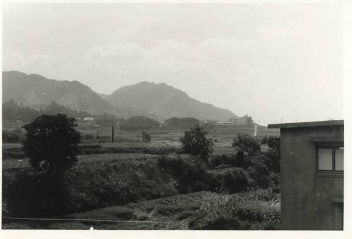 手前右に女子寮の建物、その奥に土手と田畑、その向こうに飯盛山がある昔の白黒写真