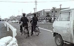 踏切の遮断機が下り、自転車に乗った2名の警察官やその後ろに停まっている自動車が電車が通過するのを待っている白黒写真