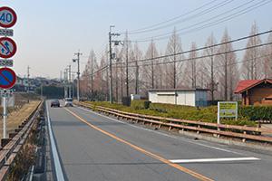 標識が設置されている道路を車が走行しており、右側には赤い屋根や白い外壁の倉庫のような建物が建っている写真