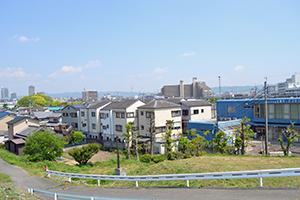 舗装された道路とガードレールできれいに整備され、ガードレールの奥にはマンション住宅が並び遠くには高層ビルが見える今の写真
