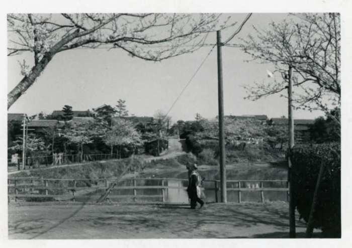 帽子を被り歩く人の横に木製の柵、その向こうに四方黒池があり、四方黒池の左側に道路が通り、住宅が広がっている昔の白黒写真