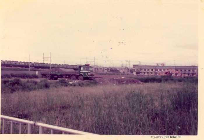 草むらにトラックやショベルカーが停まっており、後方に電車が走っている昔の緑町の写真