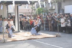 神社の一角に藁のござの上でじ膝をついて的に向かい弓を引いている写真