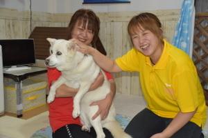 赤い洋服を着た女性が白い犬を抱っこしており、黄色い服を着た女性が犬の頭に手を置いて笑顔で写っている写真