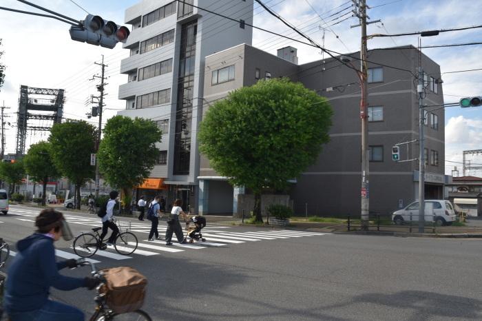 ビルが建ち並ぶ歩道に樹木が植えられており、ビルの前の横断歩道をベビーカーを押して渡る人や自転車で渡る人たちの写真