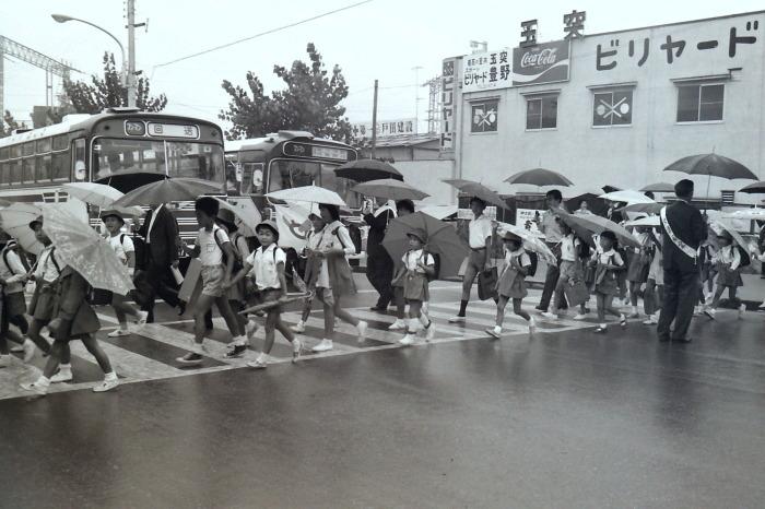 ビリヤード場前の横断歩道を傘をさした児童たちが渡っている昔の白黒写真