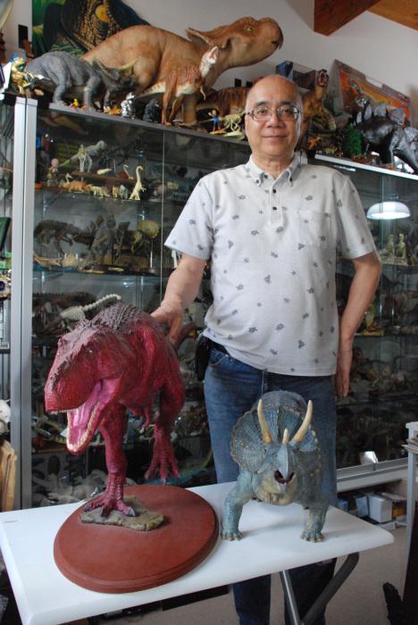 白い机上に赤色と青色の恐竜の模型が置いてある横で眼鏡をかけた荒木一成さんが立っており、後ろのガラスケースの上や中に沢山の恐竜模型がある写真