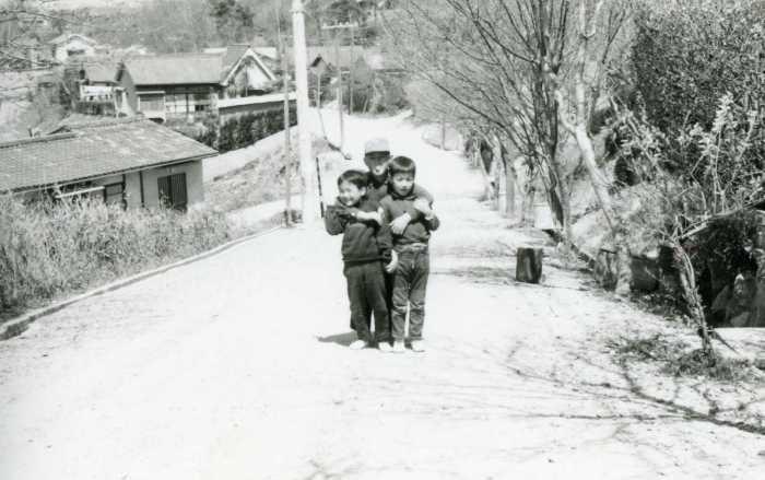 道路が舗装されていない緩やかな勾配のある道の真ん中で男の子3人が肩を組んでおり、道路の左側が一段低くなっている土地に民家が並んでいる昔の白黒写真