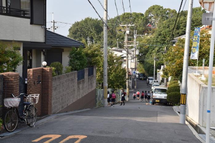 緩やかな坂道の両側が住宅街になっており、舗装された道路を歩くランドセルを背負った子供たちと車が停まっている写真