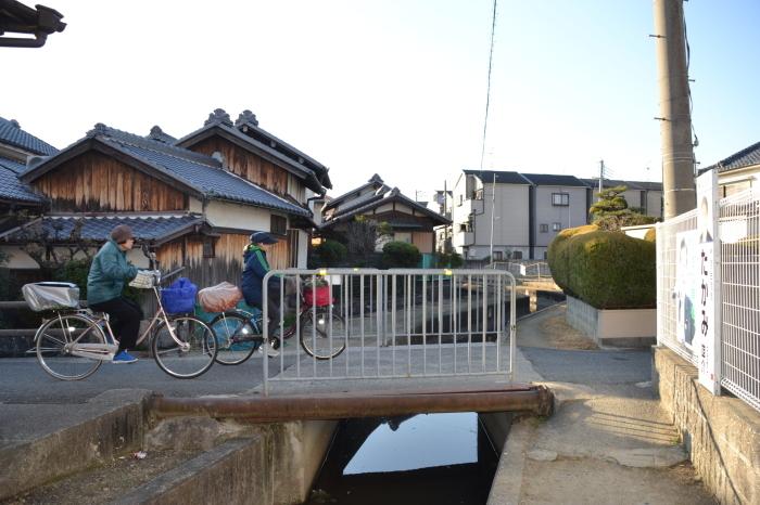 住宅街にある水路にかかる石橋に柵が設けられ、自転車のカゴと後部座席に荷物を載せた人たちが水路の上を自転車で通っている今の写真