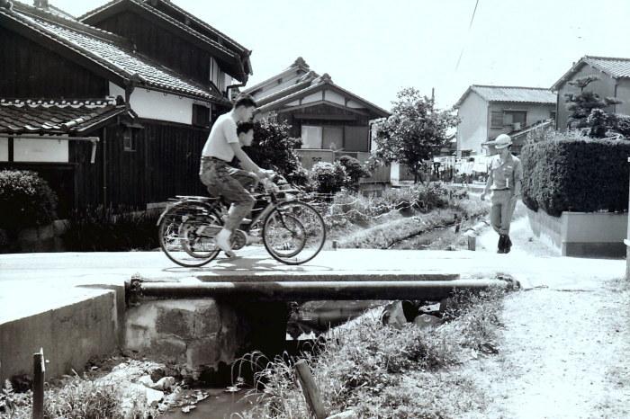 住宅街にある水路にかかる柵のない石橋を2台の自転車が渡っている昔の白黒写真