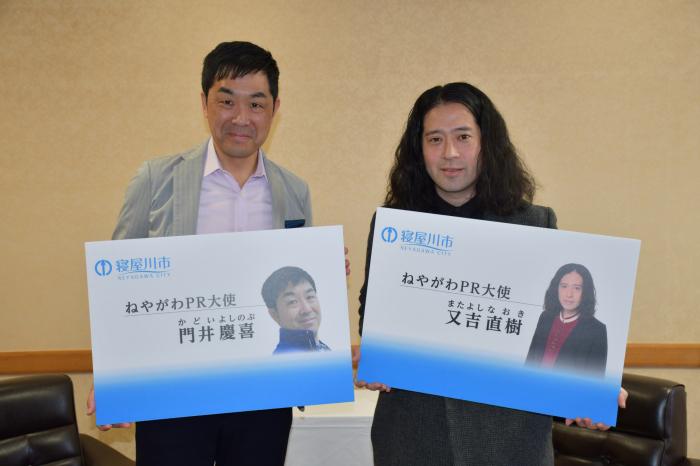 ねやがわPR大使と名前が書いてある拡大された名刺を持って記念撮影している又吉直樹さんと門井慶喜さんの写真