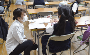 長机の席に座っている生徒と話をしている白いシャツを着た女性の写真