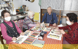 机の上に色鉛筆と画用紙を広げて、3名の女性が山口さんに色鉛筆画を習っている写真