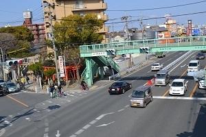 真っ直ぐに通っている道路に黄緑色の歩道橋が渡っており、歩道橋の左側には大きな木と赤い鳥居が見え、道路にはたくさんの車が走っている今の写真