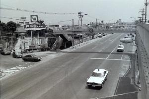 真っ直ぐに走っている道路に歩道橋が渡っており、歩道橋の左側には大きな看板、右側の車線を白い車が2台走っている昔の白黒写真