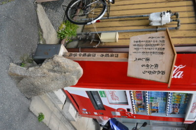 自動販売機の横に設置された、「ぬれながら旅人守る道しるべ」と書かれた木製の看板と石の道標の写真