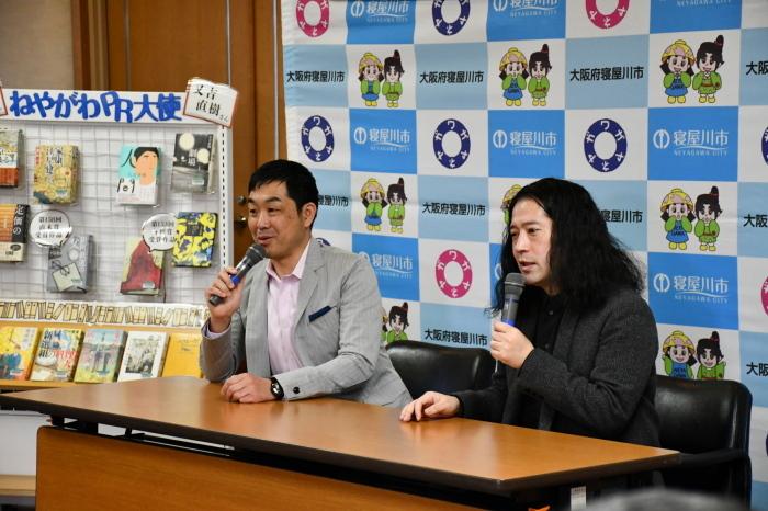ねやがわPR大使の又吉直樹さんと門井慶喜さんが長机の席に座りマイクを持ち正面を向いて話をしている写真