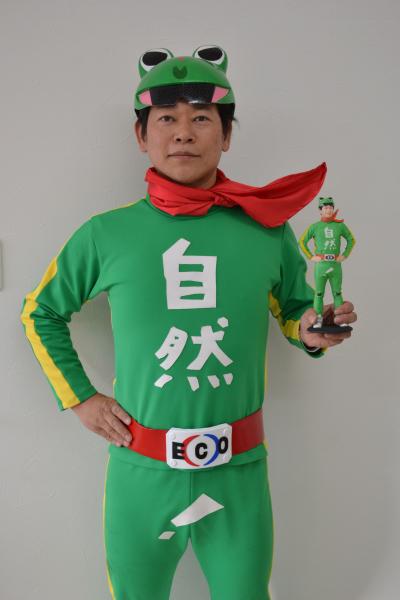自然（ECO）エコと書いてある緑色の衣装を着て、首に赤いマフラー、頭にカエルのヘルメットを被り右手は腰に当て、左手には同じ格好をしたフィギアを持っている平田彰宏さんの写真