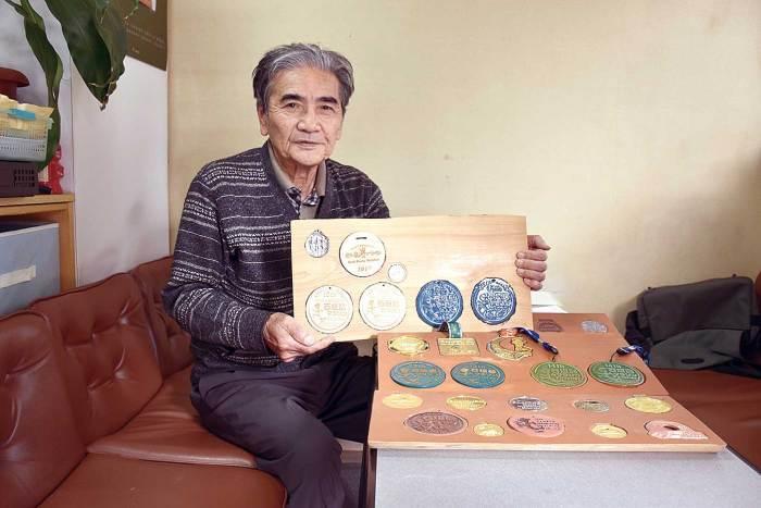 机上に置かれた板に沢山のメダルが収納されており、ソファーに座りメダルが収納された長方形の板を持っている平田宗一郎さんの写真