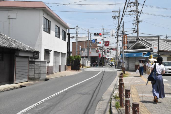 道路沿いに平屋建てや2階建ての建物が立ち並んでいる先に信号の設置された交差点があり、右側の歩道を歩く人々の写真