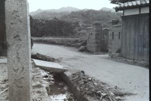 舗装されていない道の左側に記念碑が建ち、用水路が流れており、右側には木造の家が建っている白黒写真