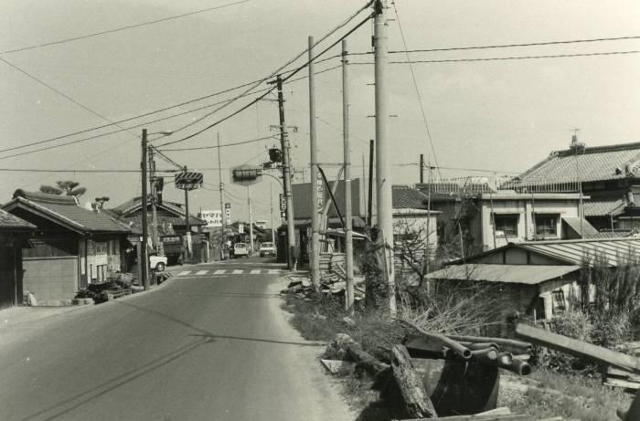 道路沿いに平屋建てが立ち並んでいる先に信号が設置された交差点がある昔の白黒写真