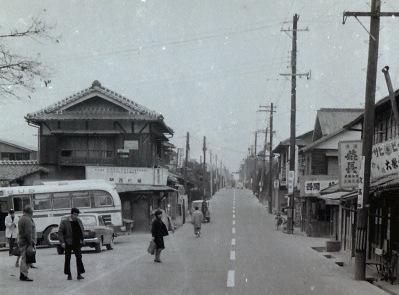 道路の両脇には商店が立ち並び、バスから乗客が降りてきている白黒写真