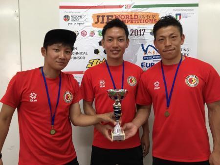 チームの赤いシャツを着てメダルを首に下げ、トロフィーに手を添えている3名の男性の写真