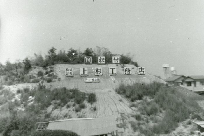 小高い丘の斜面に「国松台株式会社上杉工務店」と書かれた看板があり右奥に三井団地の給水塔2塔が写っている昔の白黒写真