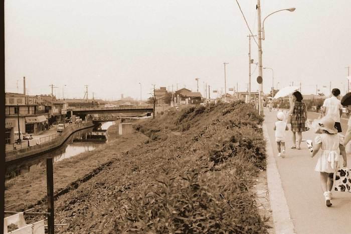 夏草が生い茂った土手沿いの道を日傘をさした親子たちが歩いている奥に橋が見える昔の白黒写真