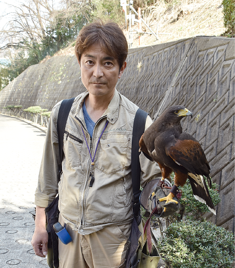 ベージュ色の作業着を着た田中和博さんのグローブをつけた手に鷹が止まっている写真