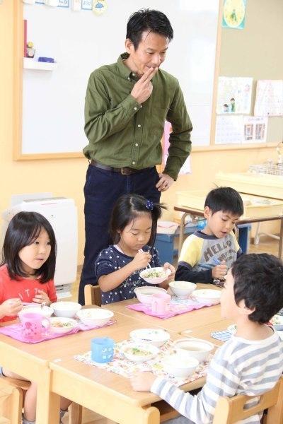 机をくっつけて向かい合わせで給食を食べている子供達とその様子を見ている田中啓昭さんの写真