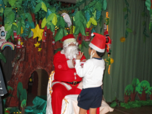 お楽しみ会にてクリスマスツリーの前で椅子に座っているサンタクロースとサンタ帽を被った男の子が話をしている写真
