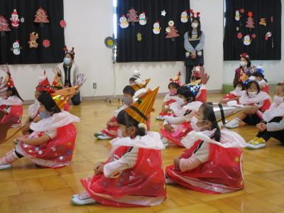 三角帽子に赤い衣装を身にまとった女の子たちと三角帽子に白いブラウスを着た男の子たちがクリスマスや雪のイラストが飾られた教室で体育座りをしている写真