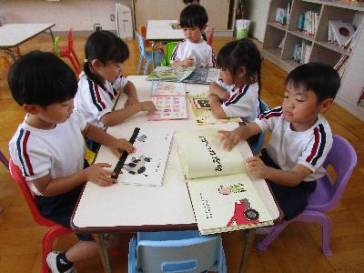 教室でテーブルごとに分かれて本を見ている子供たちとエプロン姿の先生が子供たちに教えている写真