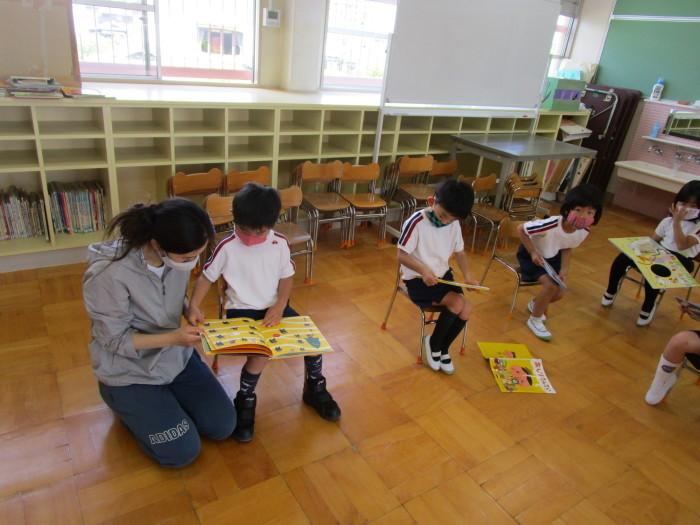 教室でパーカーを着た先生が椅子に座っている子供たちと一緒に絵本を見ている写真