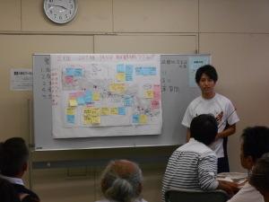 模造紙が貼られたホワイトボードの前に立って話をしている男性の写真