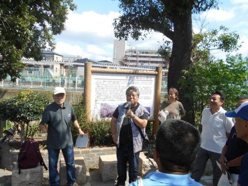 公園の案内看板の前に立って拡声器で挨拶をしている公園みどり課の代表者の男性と話を聞いている参加者の写真