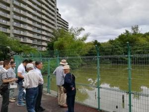 池の左側に立っている大きなマンション、池のフェンスの前に立っている見学会参加者の写真