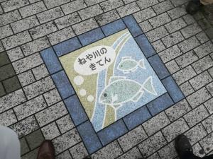 「ねや川のきてん」という文字と魚の絵が描かれた道路のタイルの写真