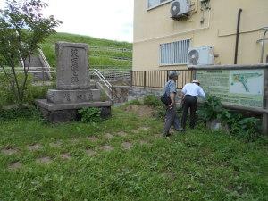 公園内に立てられた大きな石碑、その横に立っている案内看板を見ている2人の男性の写真