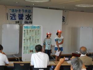 オレンジ色の帽子を被った2名の女子生徒が前に立ち活動紹介を行っている写真「