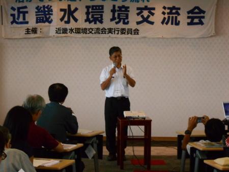 近畿水環境交流会と書かれた幕が貼られた前で、右手にマイクを持った実行委員長の澤井さんが挨拶を行っている写真