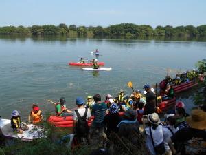 川の中央に赤色と白色のカヌーが浮かんでおり、手前の岸辺にEボートの体験を待っている参加者が並んでいる写真