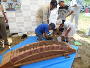 水色のシートの上でレンガアーチ橋作りを行っている3名の子どもと指導している男性の写真