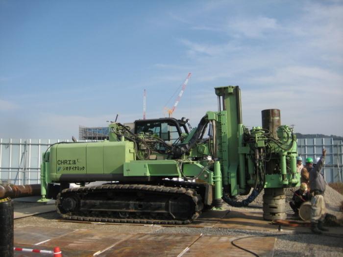 横から見た緑色の車体の先に鋼管がはめられ、右側の工事スタッフが右手を上げている写真