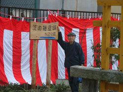 公園を隔てた柵沿いに大きな紅白幕を掛けた前で、男性が「茨田樋遺跡水辺公園」と書かれた手作りの木製の看板に右手を添えている写真