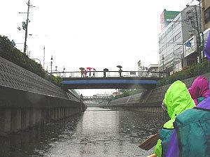 参加者がレインコートを着て寝屋川をEボートに乗り漕いでいるなか、奥の橋から見物客が傘をさして見ている写真