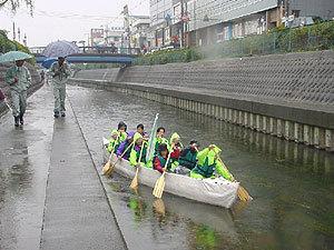 雨が降る中、レインコートを着た10名の参加者がEボートに乗り漕いでいるのを、川沿いを傘を差して歩く2名の人が見ている写真